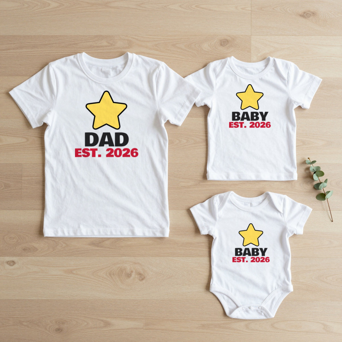 Dad Est. 2026 & Baby Est. 2026 New Dad matching set flat lay on oak floor: grown-up tee, kids tee and baby grow