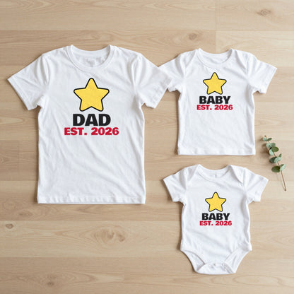Dad Est. 2026 & Baby Est. 2026 New Dad matching set flat lay on oak floor: grown-up tee, kids tee and baby grow