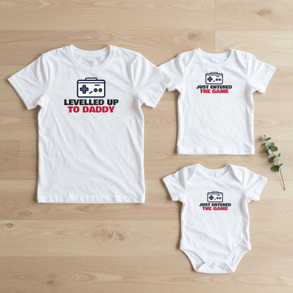 Levelled Up To Daddy & Just Entered The Game matching set flat lay on oak floor: grown-up tee, kids tee and baby grow